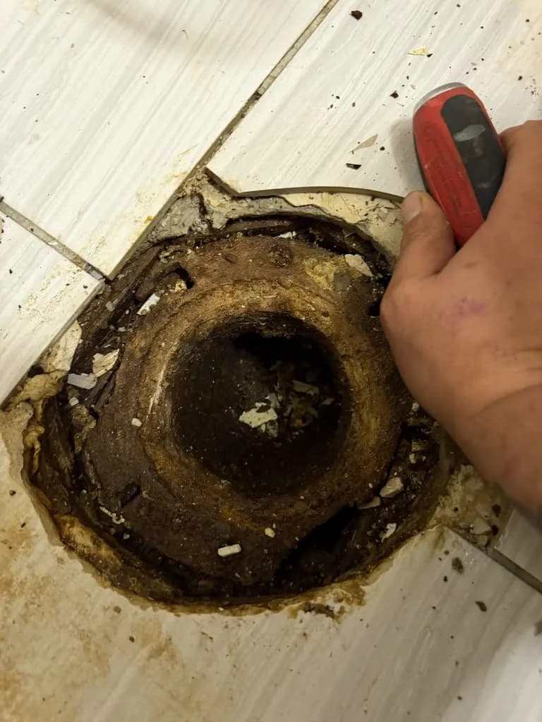 A hand using pliers to clean an old corroded toilet flange